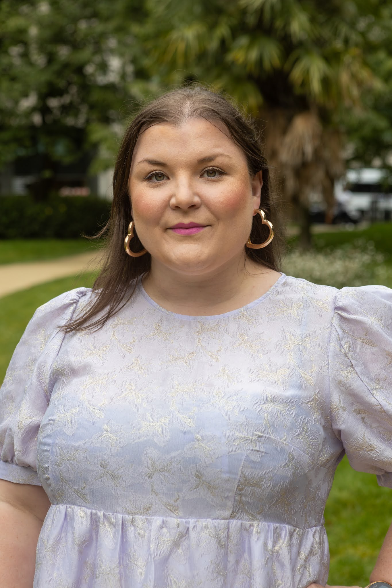 Kvinne med brunt skulderlangt hår. Med rosa leppestift og hjerte forma øreringar. Med lilla glitterkjole. Står foran eit tre og gress plen